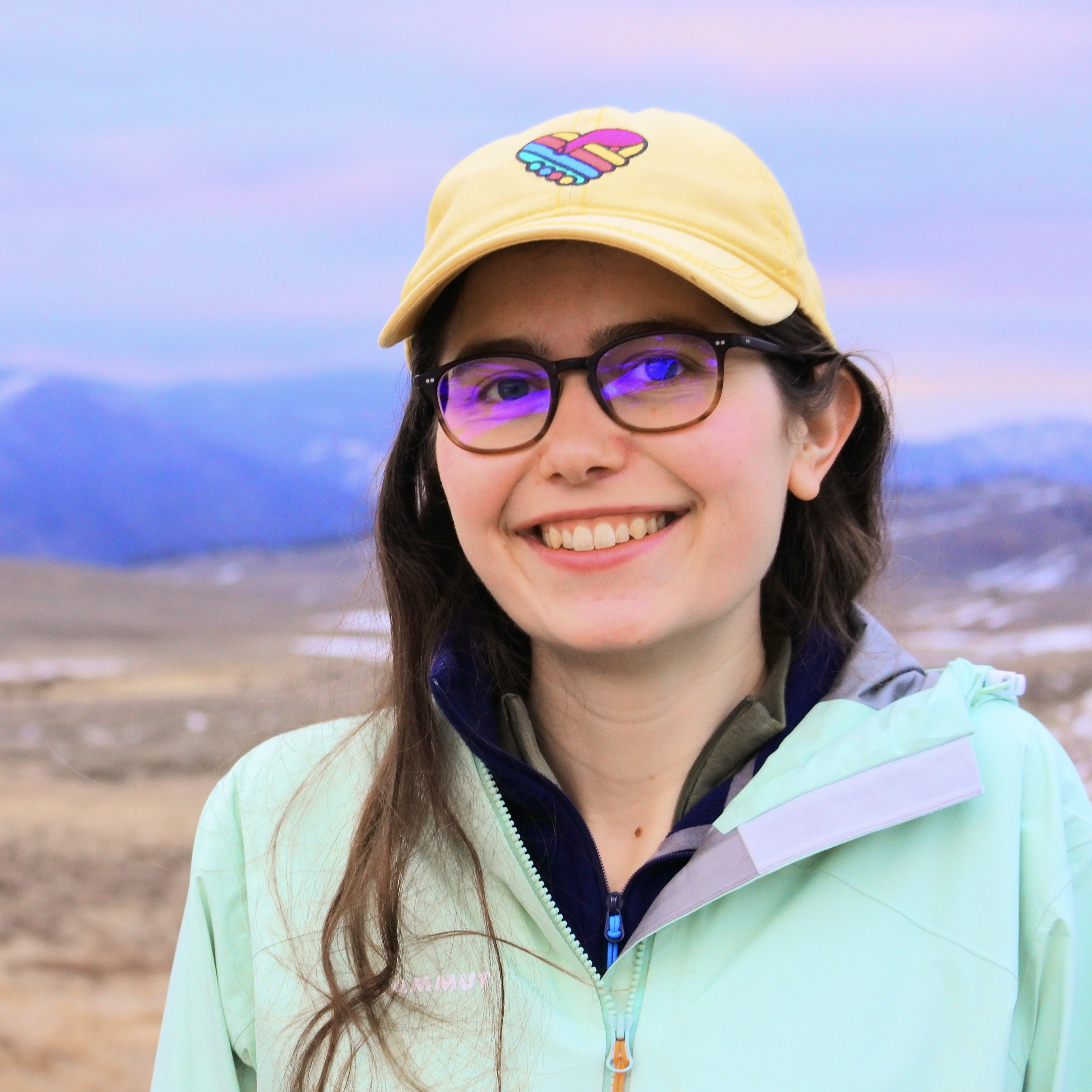 Portrait of a young smiling woman with long dark hair, glasses, and a yellow cap. In the background are the grassland and mountain chains of Yellowstone National Park.
