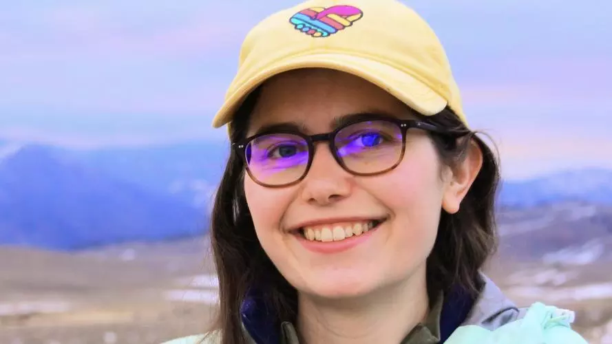 Portrait of a young smiling woman with long dark hair, glasses, and a yellow cap. In the background are the grassland and mountain chains of Yellowstone National Park.